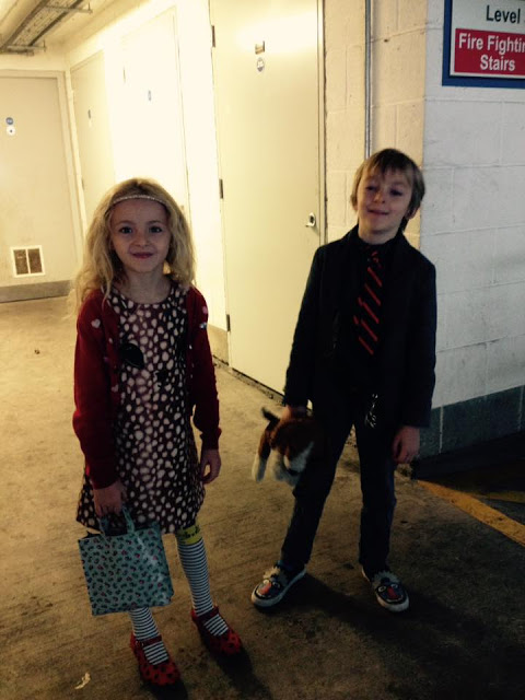 George & Molly. Molly dressed in a dress and a cardigan with gold hairband. George smiling wearing a grey suit.